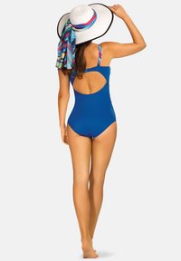 Niebieski jednoczęściowy kostium kąpielowy z wycięciem na plecach, cienkimi ramiączkami z kolorowym wzorzystym akcentem i szerokim białym kapeluszem przeciwsłonecznym z apaszką.