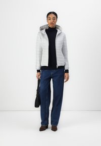 Hellgraue Pufferjacke mit horizontalen Streifen, Kapuze und Reißverschluss; kombiniert mit einem schwarzen Rollkragenpullover und blauen weit geschnittenen Jeans, braunen Schuhen.