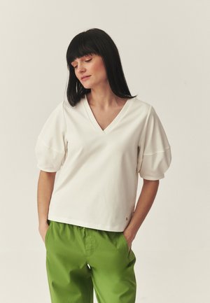 Mujer con cabello largo y negro que lleva una blusa blanca de cuello en V con mangas abullonadas y pantalones verde brillante, manos en los bolsillos, sobre un fondo liso.