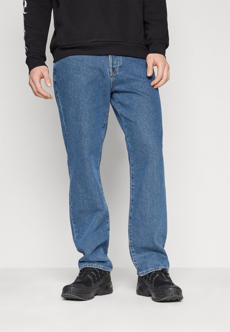 Personne portant un jean bleu coupe droite, des baskets noires et une chemise noire à manches longues, debout devant un fond uni.
