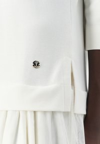 Maglia bianca lavorata a maglia con scollatura rotonda e maniche corte, dotata di un emblema decorativo nero e argento all'orlo e di spacchi laterali.