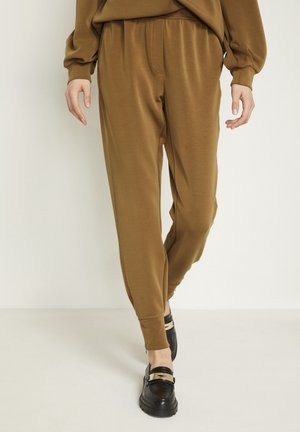 Personne portant un pantalon jogger marron fuselé et des mocassins noirs marchant vers l'avant sur un fond uni.