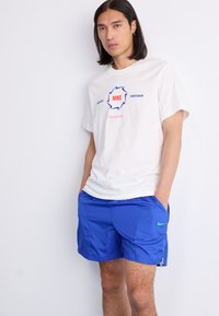 Hvid bomulds t-shirt med "Nike" og "Sportswear" trykt i blåt og rødt. Blå shorts med et grønt Nike-logo og sidelommer.