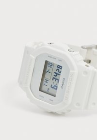 Montre numérique Casio G-Shock blanche avec un écran rectangulaire, un bracelet en caoutchouc, et un texte indiquant l'heure et les fonctions, affichant des chiffres clairement.