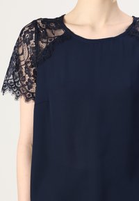 Mornarska bluza s kratkimi rokavi, ki ima zapleten črn čipkast rob na ramenih. Gladka tkanina z okroglim izrezom in sproščenim krojem.