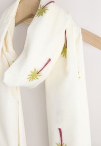 Écharpe de couleur crème avec broderie florale verte et rouge suspendue sur un cintre en bois sur fond blanc.
