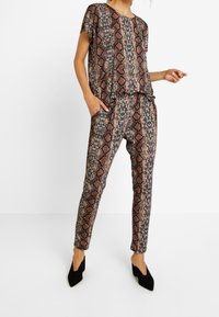 Femme portant un ensemble coordonné avec haut et pantalon à imprimé serpent, se tenant avec une main dans la poche et des chaussures à talons noirs sur un fond blanc.