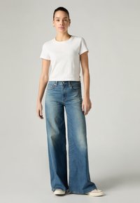 Λευκό κοντομάνικο t-shirt σε συνδυασμό με φαρδιά τζιν παντελόνια σε ανοιχτό μπλε χρώμα. Τα τζιν έχουν ξεβαμμένο φινίρισμα και μπροστινές τσέπες, ολοκληρωμένα με λευκά αθλητικά παπούτσια.