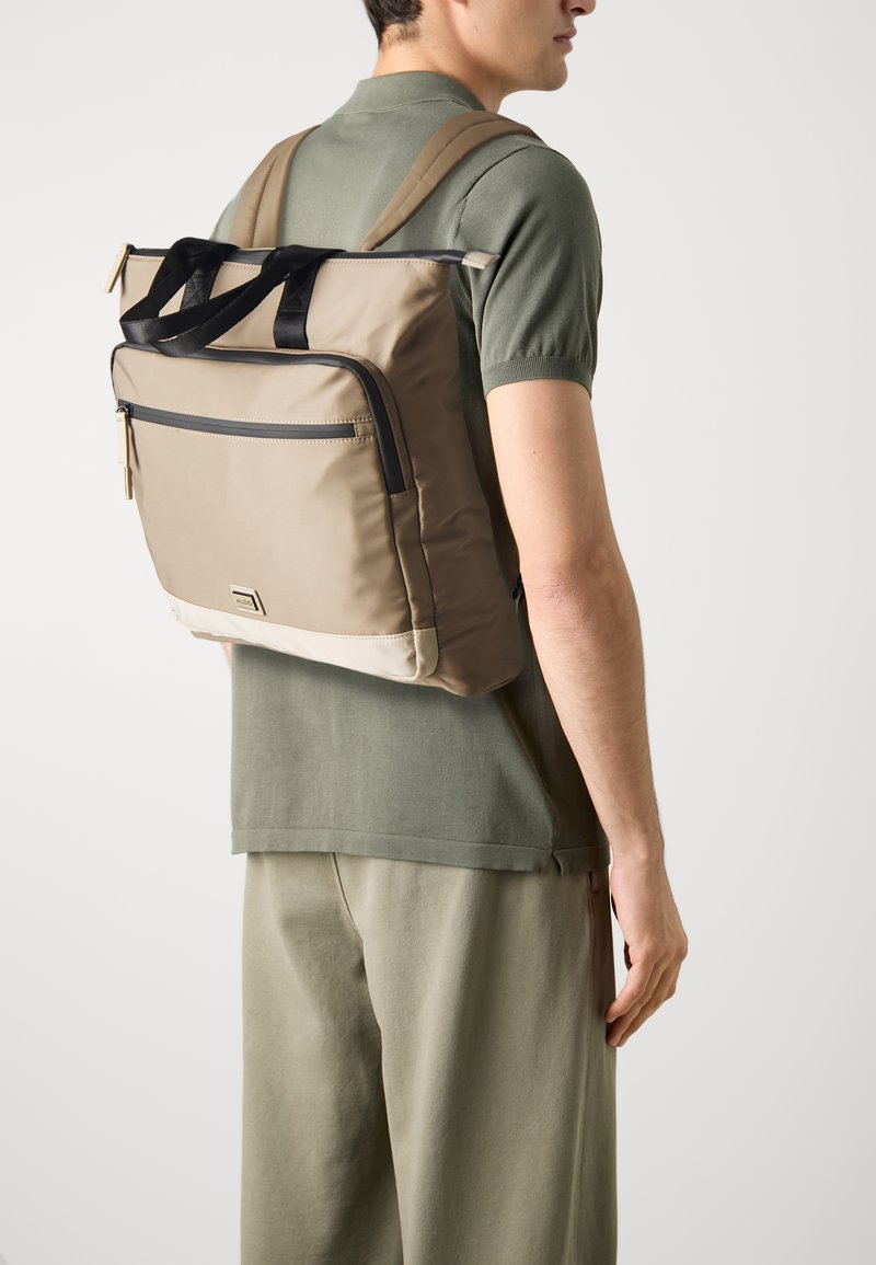 Beige gewatteerde rugzak met zwarte accenten, voorzien van een voorvak met rits en dubbele schouderbanden, gedragen over een groene top en grijze broek.