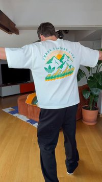 Osoba stojąca z wyciągniętymi ramionami, ubrana w białą koszulkę z zielono-żółtą grafiką gór i napisem "adidas" na plecach.