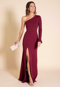 Vestido burdeos con un escote asimétrico, manga larga ajustada y una alta abertura lateral, confeccionado con una tela suave. Los accesorios incluyen un clutch transparente.