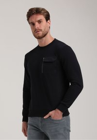 Laikkuun sweatshirt sinisen värisenä, jossa on pitkät hihat, pyöreä kaula-aukko ja vetoketjullinen rintatasku. Ribbattu hihansuu ja helma. Yhdistetty vaaleanharmaisiin farkkuihin.