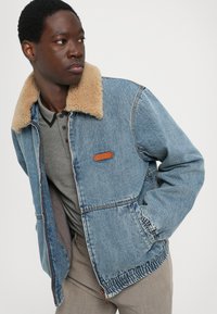 Denim müts, millel on beež faux fur krae, sinine pesu, elastsed mansetid ja küljetaskud. Rinnal on nahklabel.