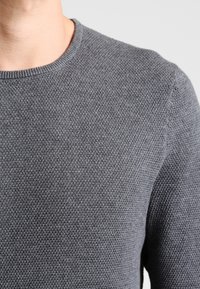 Šedý pletený svetr s kulatým výstřihem, vybavený strukturovanou tkaninou a žebrovanými manžetami, těsně přiléhající k tělu, bez viditelných vzorů nebo kovových doplňků.