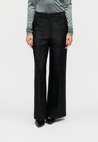 Μαύρο παντελόνι φαρδύ με λεία υφή, πιέτες μπροστά και θηλιές για ζώνη, συνδυασμένο με ανοιχτοπράσινο μπλουζάκι μακριάς μανίκας και μαύρα παπούτσια.