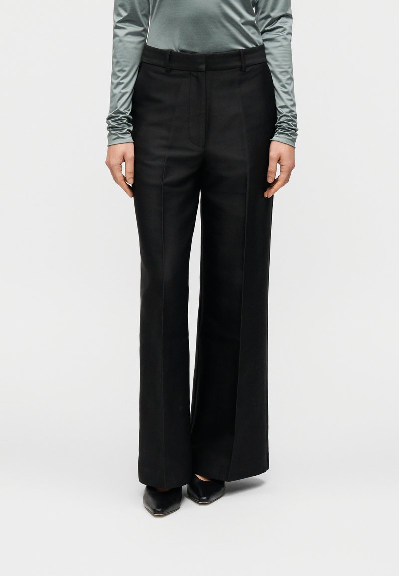 Μαύρο παντελόνι φαρδύ με λεία υφή, πιέτες μπροστά και θηλιές για ζώνη, συνδυασμένο με ανοιχτοπράσινο μπλουζάκι μακριάς μανίκας και μαύρα παπούτσια.