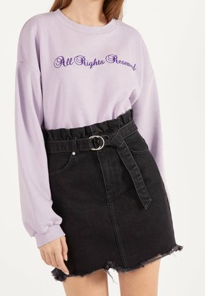 Kvinde iført en lyse lilla sweatshirt med teksten "All Rights Reserved" og et sort, højtaljet denimskørt med bælte og frynset kant.