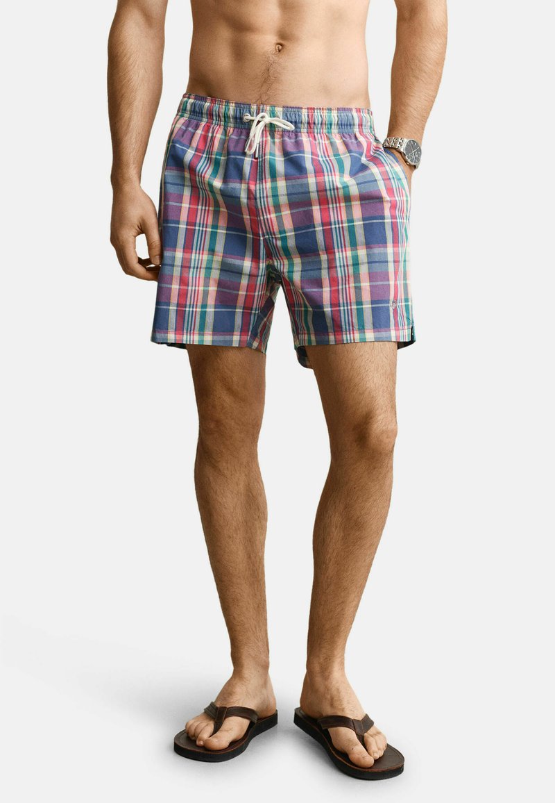 Homme portant un short de bain à carreaux multicolores avec cordon de serrage, une montre au poignet gauche et des tongs marron foncé aux pieds nus.