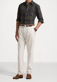 Camicia a quadri verde e blu, pantaloni bianchi plissettati, cintura in pelle marrone e mocassini marroni. Orologio al polso, in posa in piedi.