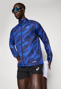 Blåmønstret løbejakke med høj krave, lange ærmer og lynlås foran. Sorte shorts og reflekterende detaljer fuldender det atletiske look.