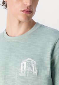 Šviesiai žalia, trumpomis rankovėmis marškinėliai, pagaminti iš minkštos medžiagos, su balta siuvinėta architektūriniu raštu ant krūtinės.