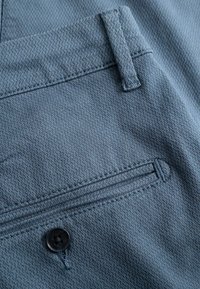 Mėlynos tekstūruotos kelnės su nugarine kišene, kurią puošia saga ir tvarkingi siūlai, didinantys patvarumą ir konstrukciją.