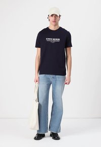 Mørkeblå bomulds T-shirt med "TOMMY HILFIGER" logo, parret med lyseblå baggy jeans og sorte sko; holder en hvid tote bag.