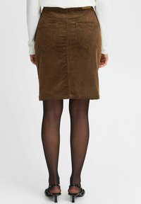 Jupe en velours côtelé marron à coupe droite, avec poches arrière et ourlet au-dessus du genou, associée à des sandales à talons noires et des collants transparents.