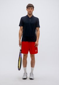 Μαύρο μπλουζάκι πόλο με κοντά μανίκια, κόκκινα σορτς, λευκές κάλτσες και γκρι αθλητικά παπούτσια. Κρατά ρακέτα τένις. Απλός σχεδιασμός, αθλητική εφαρμογή.