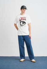 Camiseta branca com gráfico de olho e texto vermelho "OBEY", combinada com jeans azuis largos e tênis em tom de creme. Boné preto com logótipo.