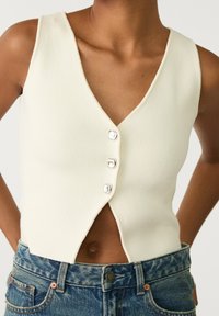Närbild av en person som bär en ärmlös krämfärgad blus med tre knappar och blå denimjeans, med händerna vilande på höfterna.