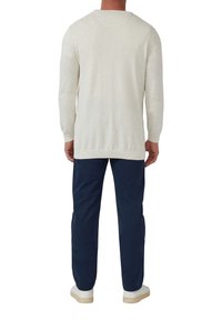 Mann, der mit dem Rücken zur Kamera steht, trägt einen hellbeigen Langarmpullover, dunkle blaue Hosen und weiße Sneakers auf einem weißen Hintergrund.