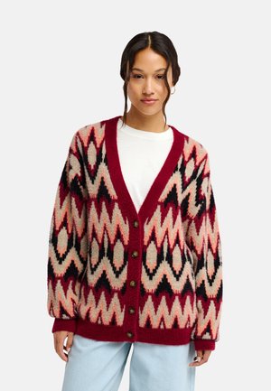 Vrouw draagt een rode, roze, zwarte en beige cardigan met zigzagpatroon en knopen over een wit overhemd en lichtblauwe broek, staand in een ontspannen houding.