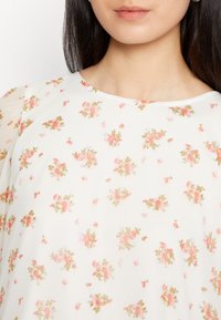 Kvinna som bär en vit blus med små rosa och gröna blommönster, synlig från halsen till övre bröstet.