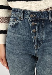 Denim jeans in faded blue with a high waist, featuring five-button closure, two front pockets, and diagonal stitching accents.