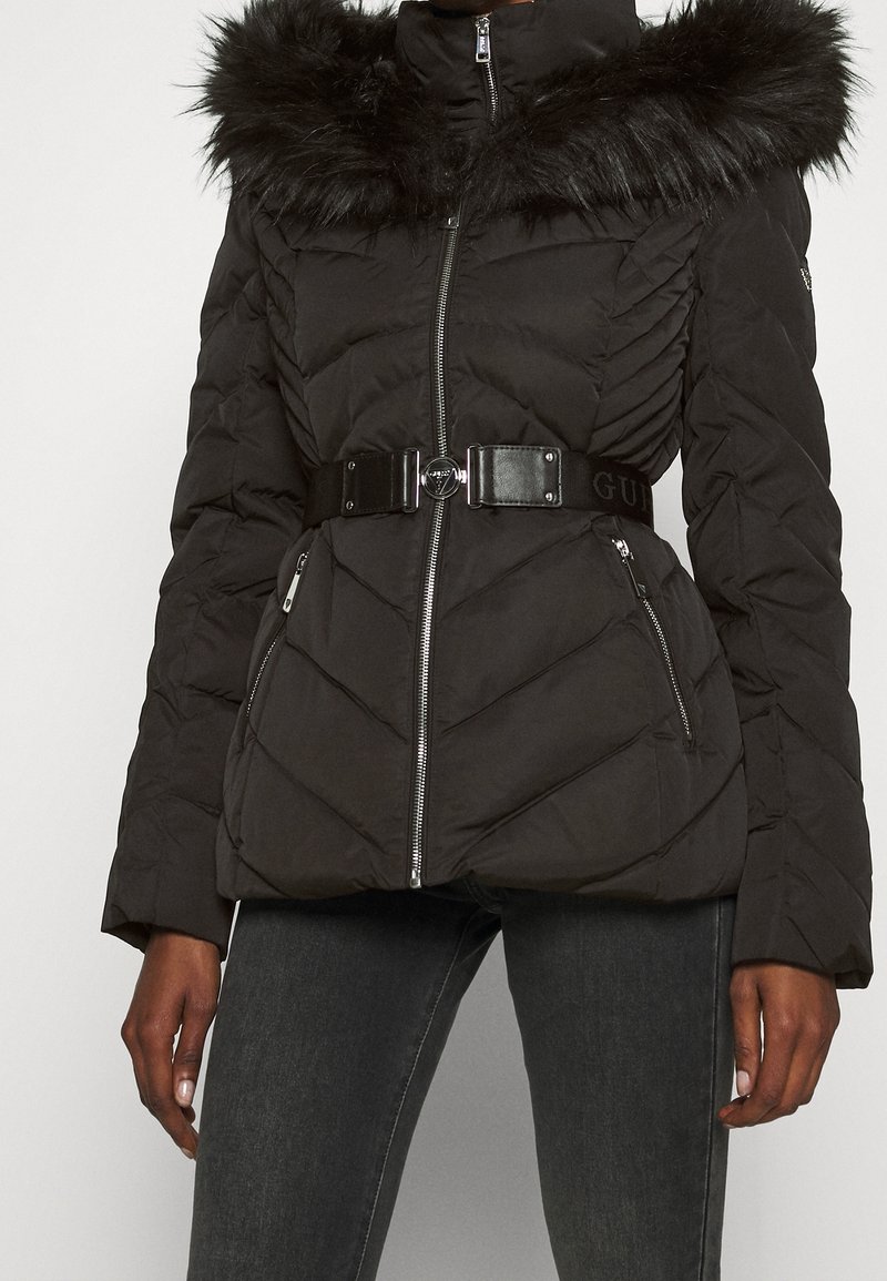 Sort pufferjakke med fuskepelskrave, chevron-quiltet design, lynlåsfortil, to sidelommer og et bredt sort bælte som accent.