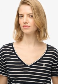 Granatowo-kremowy t-shirt w paski z dekoltem w serek i krótkimi rękawami, z małym haftowanym logo na piersi oraz miękką fakturą materiału.