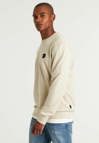 Sudadera de cuello redondo beige con acabado texturizado, que presenta un pequeño parche de logo negro en el pecho y puños acanalados en las mangas.