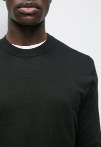 Close-up de uma pessoa a usar um suéter preto de gola redonda sobre uma camisa branca, contra um fundo claro e liso.