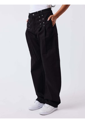 Personne portant un pantalon noir ample avec des boutons sur le devant et des baskets blanches, debout les mains dans les poches devant un fond uni.