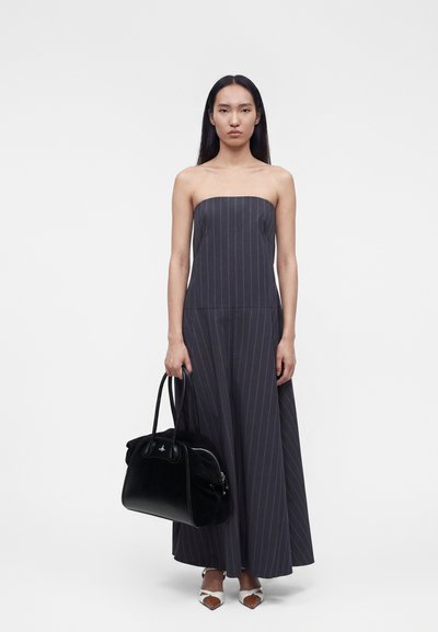 Γυναίκα με μακριά σκούρα μαλλιά, που φοράει σκούρο γκρι φόρεμα χωρίς τιράντες με ριγέ σχέδιο, κρατώντας μια μεγάλη μαύρη τσάντα, στέκεται σε λευκό φόντο.