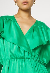 Πράσινο σατέν φόρεμα με βαθύ V-λαιμό, ρυτιδωμένο λαιμό και πτυχωτή μέση για μια φουσκωτή σιλουέτα. Λείο υφή με γυαλιστερό φινίρισμα.