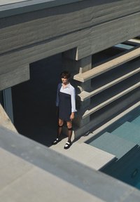 Le modèle porte une robe marine sur une chemise blanche, avec des chaussettes noires jusqu'aux genoux et des chaussures noires. Des caractéristiques architecturales en béton sont visibles en arrière-plan.