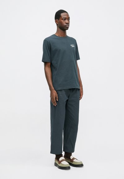 Dunkelgrünes Baumwoll-T-Shirt mit kurzen Ärmeln, kombiniert mit weit geschnittenen, dunkelgrauen Hosen und beigen Lederschuhen mit braunen Akzenten.
