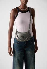 Borsa a spalla in pelle grigia con un accento in catena, hardware a forma di diamante e design curvo. Il modello indossa una canotta chiara e jeans a gamba larga.