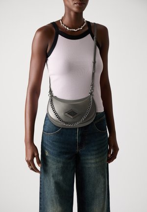 Grijze leren schoudertas met een kettingaccent, ruitvormige hardware en een gebogen ontwerp. Model draagt een lichte tanktop en wijde jeans.