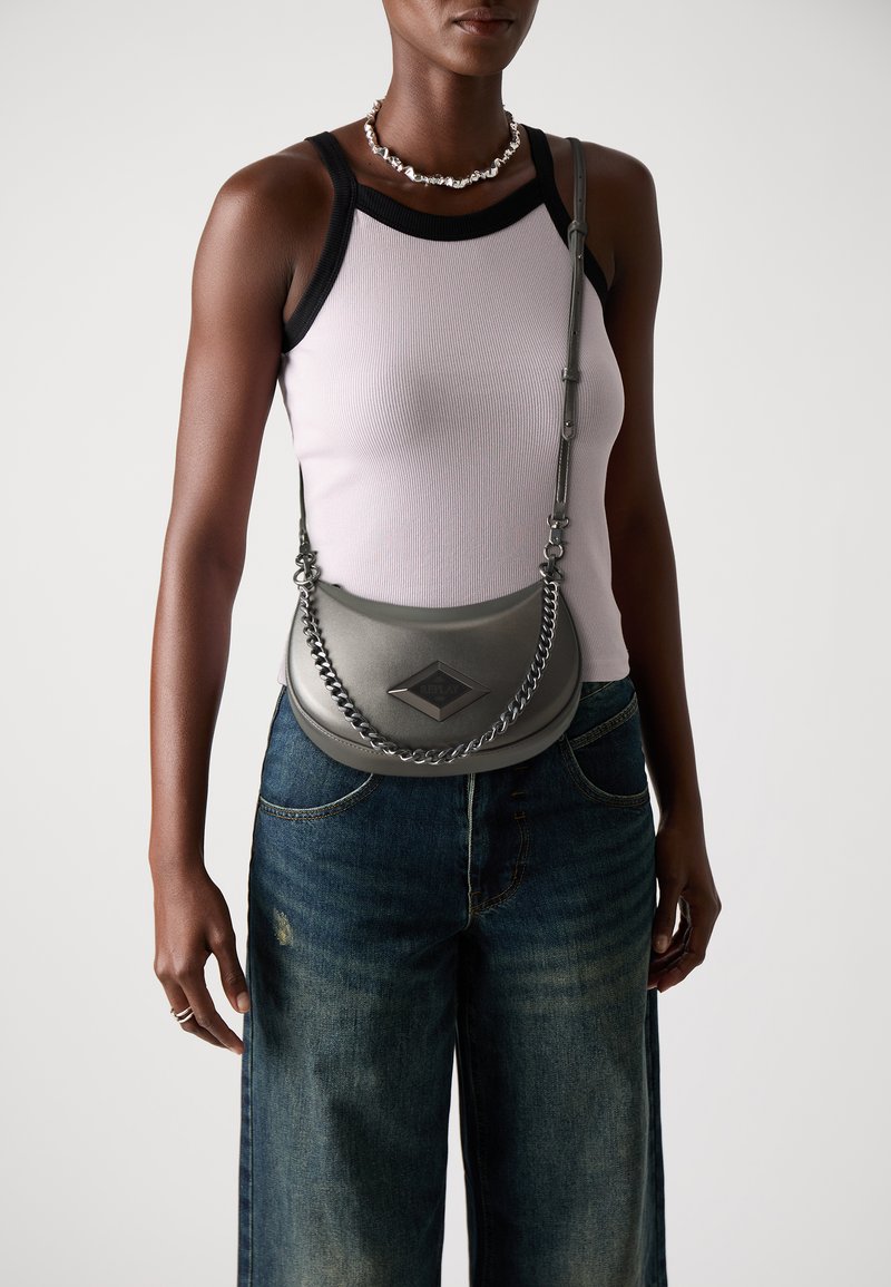 Borsa a spalla in pelle grigia con un accento in catena, hardware a forma di diamante e design curvo. Il modello indossa una canotta chiara e jeans a gamba larga.