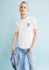 Weißes Baumwoll-T-Shirt mit einem blauen Sonnenmotiv auf der linken Brust, kombiniert mit hellblauen Jeans und einer Jeansjacke in der Hand.