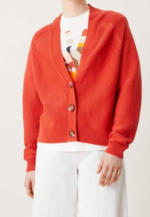 Cardigan rouge côtelé avec un décolleté en V profond, comportant quatre boutons marron sur le devant. Porté sur un t-shirt graphique blanc et un pantalon clair.