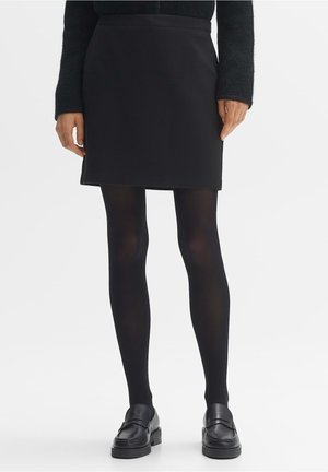 Persoon die een zwarte minirok, zwarte panty's, zwarte loafers en een donker top met lange mouwen draagt, staand tegen een effen achtergrond.